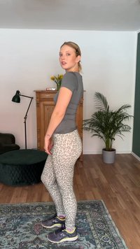 Noor naine halli pusa ja leopardimustriliste retuusidega seisab külje peal mustrilisel vaibal elutoas, kus on taimi ja puidust kapp.