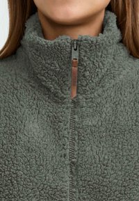 Veste en polaire zippée de couleur vert foncé, avec un col haut, un tissu texturé et une tirette de fermeture éclair accentuée en cuir avec détail de logo.