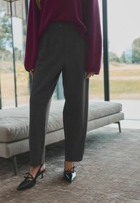 Grijze high-waisted broeken met voorplooien, gecombineerd met zwarte puntige slingback hakken met strikdetail. Gezellige wijnrode trui.