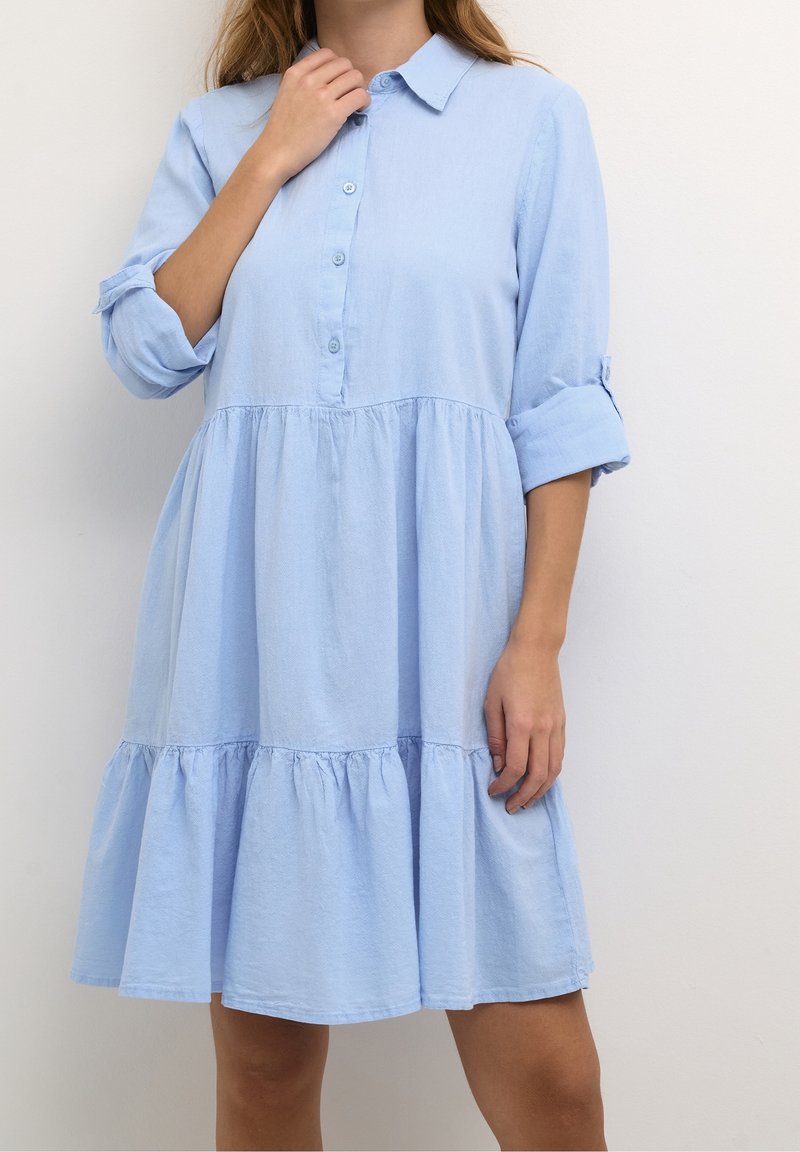 Nainen, joka on pukeutunut polvimittaisiin, vaaleansinisiin nappilistapaitaan, jossa hihat on käännetty ylös, seisoo tasaisella valkoisella taustalla.