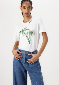 Witte t-shirt met een groen, gefotografeerde palmboomdesign; gecombineerd met hoge, blauwe denim jeans met kleine witte stippen.