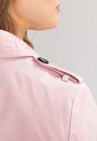 Ljusrosa trenchcoat med en slät yta, utrustad med en knappad axelrem och en krage, som visar en modern design.