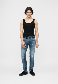 Νεαρό άτομο στέκεται με το βλέμμα προς τα εμπρός, φορώντας μαύρο αμάνικο, σκισμένο μπλε τζιν με μαύρη ζώνη με κρίκους και μαύρα αθλητικά παπούτσια.