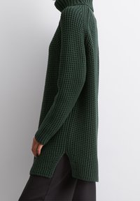 Grüner Strickpullover mit hohem Kragen, gerippter Textur und seitlichen Schlitzen am Saum. Lange Ärmel, lockere Passform und grob gestricktes Design.