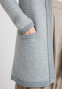 Ljusblå stickad cardigan med ett texturerat mönster, som har en synlig ficka och ribbade kanter. Tyget har ett mjukt, mysigt utseende.