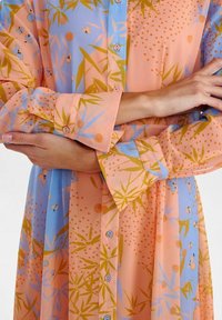 Chemise longue à boutons en tissu léger. Présente des motifs floraux roses et bleus avec des accents verts. Poignets à boutons et col décontracté.