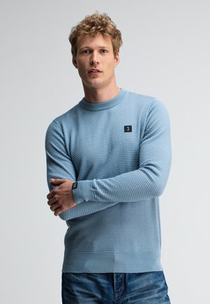 Hellblauer strukturierter Pullover mit Rundhalsausschnitt, ausgestattet mit einem kleinen Markenlogo auf der Brust und gerippten Bündchen, kombiniert mit blauen Jeans.