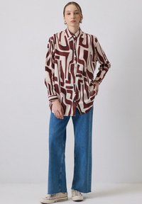 Langærmet skjorte med knaplukning i cremefarvet bund og brune abstrakte mønstre, parret med løse blå jeans og hvide sneakers.