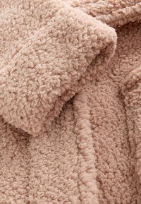 Fleece tæppe i lys pink, med en tekstureret, fluffy overflade og en blød, plysset fornemmelse samt syede kanter.