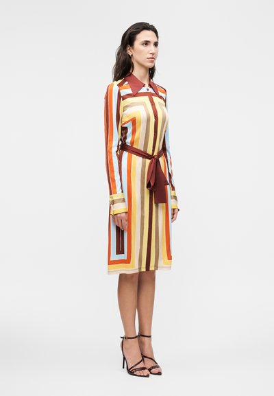 Γυναίκα που φοράει ένα πολύχρωμο ριγέ φόρεμα μέχρι το γόνατο με μακριά μανίκια, καφέ γιακά και ζώνη στη μέση, συνδυασμένο με μαύρα πέδιλα με λουράκια.