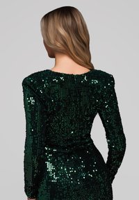 Robe verte à sequins avec manches longues et dos en décolleté arrondi. Le tissu reflète la lumière, créant un effet scintillant sur toute la surface.