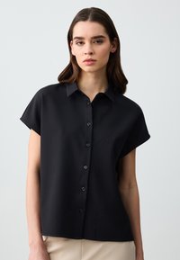 Camicia nera a maniche corte con colletto, realizzata in tessuto morbido, con una vestibilità rilassata e una leggera lucentezza.