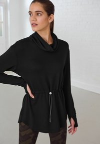 Top noir à manches longues avec un col drapé, taille à cordon coulissant et texture lisse. Présente des fentes latérales et des poignets superposés pour plus de liberté de mouvement.