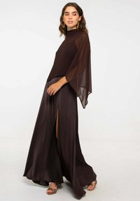 Vestido largo de color marrón oscuro con una silueta fluida, que presenta un cuello alto y mangas anchas. La abertura vertical en la falda realza el movimiento.