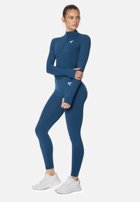 Conjunto deportivo azul con una camiseta ajustada de manga larga y leggings de tiro alto. Tejido suave con un ligero brillo, cuenta con un cierre con cremallera y un logo.