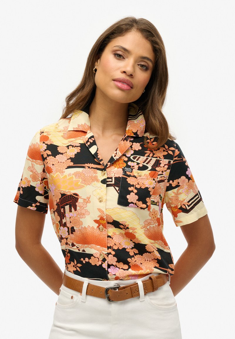 Camisa de manga corta con un patrón floral en naranja, rosa y negro sobre un fondo claro; incluye un cuello y un bolsillo.