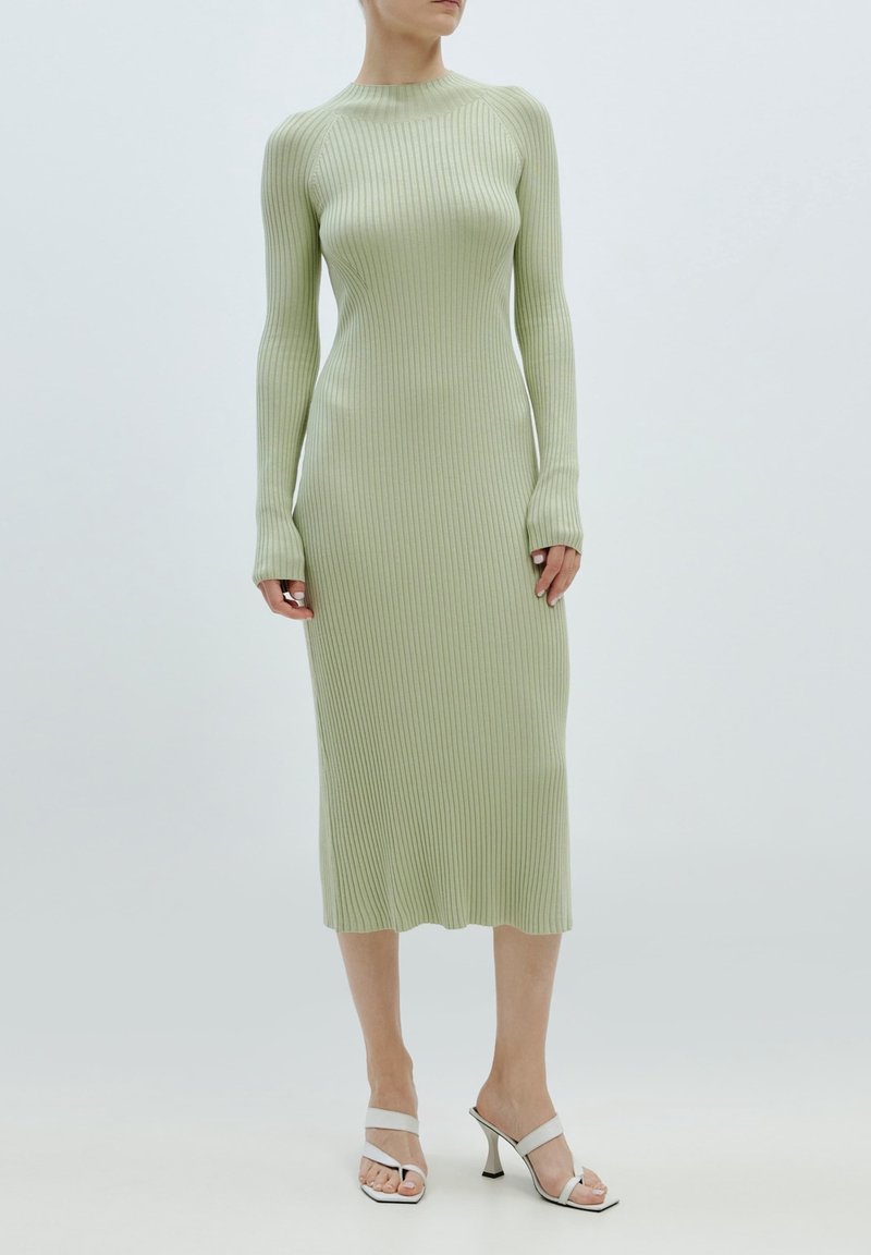 Robe à manches longues en maille côtes vert clair, silhouette ajustée, col rond, tricot texturé. Portée avec des talons blancs ouverts, design minimaliste.