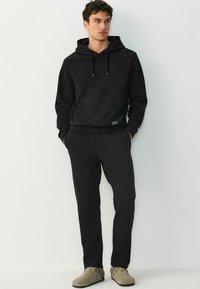 Hanorac negru cu glugă, cu textură canelată, buzunar frontal și șnururi, asortat cu pantaloni jogger negri. Încălțămintea bej completează ținuta.