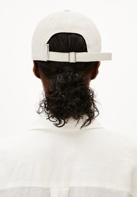 Witte honkbalpet met een gebogen klep, verstelbare band aan de achterkant en een textiele stof. Haar is gestyled in losse krullen.