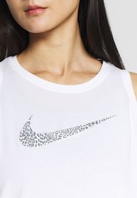Biały top na ramiączkach z szarym wzorem w panterę oraz logo Nike w kształcie szuwa na przodzie. Materiał wydaje się miękki i przewiewny, z okrągłym dekoltem.