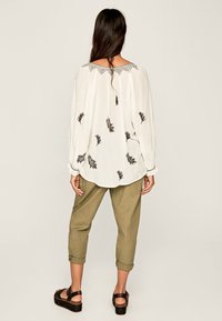 Lockere, weiße Bluse mit schwarzer Blätterstickerei, einem gewellten Ausschnitt und langen Ärmeln. Kombiniert mit gerollten olivgrünen Hosen und schwarzen Plattform-Sandalen.