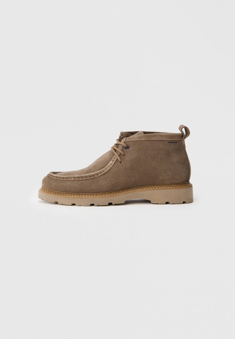 Brune ruskind ankelstøvle med beige gummisål og snørebånd foran, set fra siden på en ensfarvet hvid baggrund.