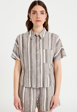 Giovane donna che indossa una camicia beige e grigia a righe verticali con bottoni e pantaloncini abbinati, in piedi davanti a uno sfondo bianco uniforme.