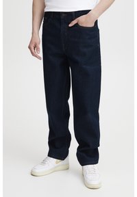 Vaqueros de mezclilla azul oscuro con un ajuste relajado, presentan costuras naranjas, cinco bolsillos y bragueta con botones. Estilizados con zapatillas blancas.