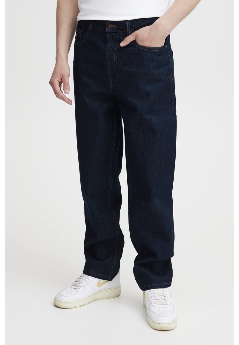 Vaqueros de mezclilla azul oscuro con un ajuste relajado, presentan costuras naranjas, cinco bolsillos y bragueta con botones. Estilizados con zapatillas blancas.