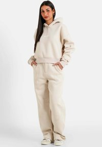 Hoodie cropped beige con tasca frontale e pantaloni morbidi a gamba larga abbinati in vita alta. Il tessuto appare morbido e strutturato, con dettagli di design minimi.