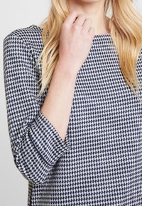 Marineblå och vit hundtandmönstrad topp med trekvartsärmar, som har en rund halsringning och subtil kontrasterande kant på ärmarna.