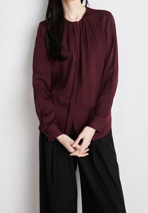Femme aux longs cheveux foncés portant un chemisier bordeaux à manches longues et un pantalon large noir sur un fond clair uni.