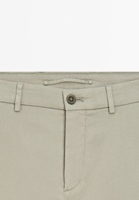 Ljusgrå byxor med en slät textur, som har knappstängning, bältesöglor och en framficka. Sömnaden är neat och jämn.