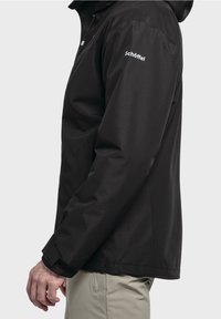 Muž v černé voděodolné bundě Schöffel a béžových kalhotách, zobrazený z boku na jednoduchém světlém pozadí.