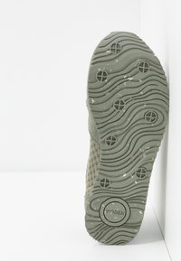 Hellgrüner Sneaker mit einer strukturierten Netz-Oberfläche, runder Gummisohle mit wellenförmigem Muster und rundem Markenlogo in der Nähe der Ferse.