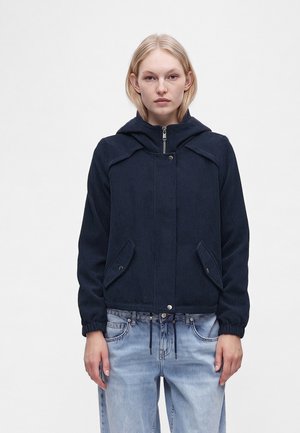 Ung kvinde iført en marineblå hættetrøjejakke med forlommer og lyseblå falmede jeans, der står foran en ensfarvet baggrund.