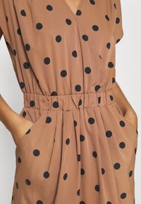 Ruskea, lyhythihainen mekko, jossa on V-kaula-aukko, elastinen vyötärö ja suuria mustia pilkkuja; sisältää sivutaskut ja plisseen yksityiskohtia.