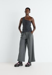 Top grigio scuro a spalla unica con bottoni, abbinato a pantaloni grigi a gamba larga. Il modello indossa scarpe nere robuste con un accento rosa.