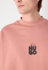 Rózsaszín, magas galléros pulóver, amelyen egy fekete, torzított "HUGO" logó található. Puha, textúrált anyagból készült, laza szabással.