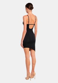 Schwarzes, figurbetontes Kleid mit Spaghetti-Trägern, offener Rückenpartie und asymmetrischem Saum. Glatte Textur und figurbetonter Schnitt. Ein Paar nude-farbene High Heels.