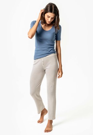 Frau barfuß, trägt ein blaues Kurzarmoberteil und beige gestreifte Hosen, steht und blickt nach unten, wobei sie mit der Hand ihr Haar berührt.