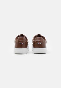 Baskets en cuir marron avec semelle en caoutchouc blanche, présentant un design arrondi et un accent tricolore à l'arrière. Texture lisse et style minimal.