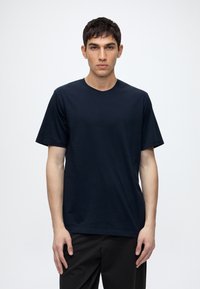 Young man with short dark hair wearing a plain dark blue short-sleeve t-shirt and black pants, standing against a white background.