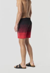 Homme torse nu debout, portant un short de bain dégradé du rouge au noir et des sandales noires à deux brides, vu de dos et de profil sur un fond uni.