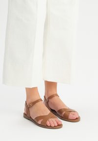 Brązowe skórzane sandały mają krzyżujące się paski i zapięcie na kostce. Projekt jest płaski z teksturowaną gumową podeszwą. Palce są widoczne.