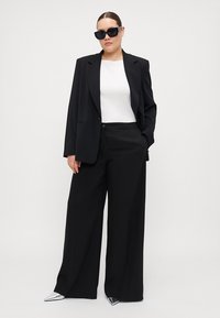 Svart drakt med skreddersydd blazer og vide bukser, kombinert med en hvit topp og sølvfargede spisse sko, komplettert med svarte solbriller.