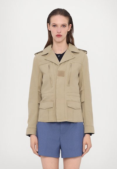 Veste kaki coupée avec épaulettes, plusieurs poches zippées et à rabat, et une finition texturée. Associée à un short bleu.