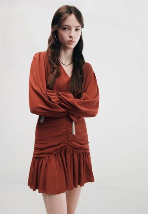Jeune femme aux longs cheveux bruns portant une robe froncée couleur rouille, debout les bras croisés devant un fond uni.