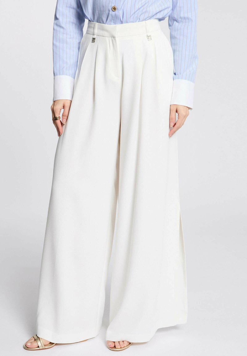 Pantaloni bianchi a gamba larga con pince in vita e spacco laterale. Il modello li abbina a una camicia a righe blu e bianche e sandali dorati.