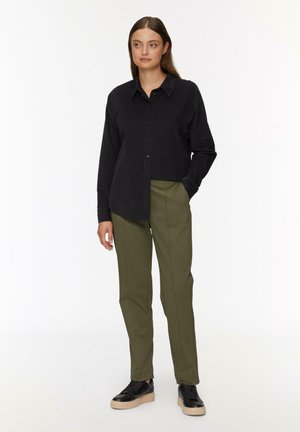 Schwarzes Hemd aus Cordstoff, kombiniert mit olivgrünen Hosen. Das Outfit hat einen lässigen Schnitt und schwarze Sneaker mit einer klobigen Sohle.